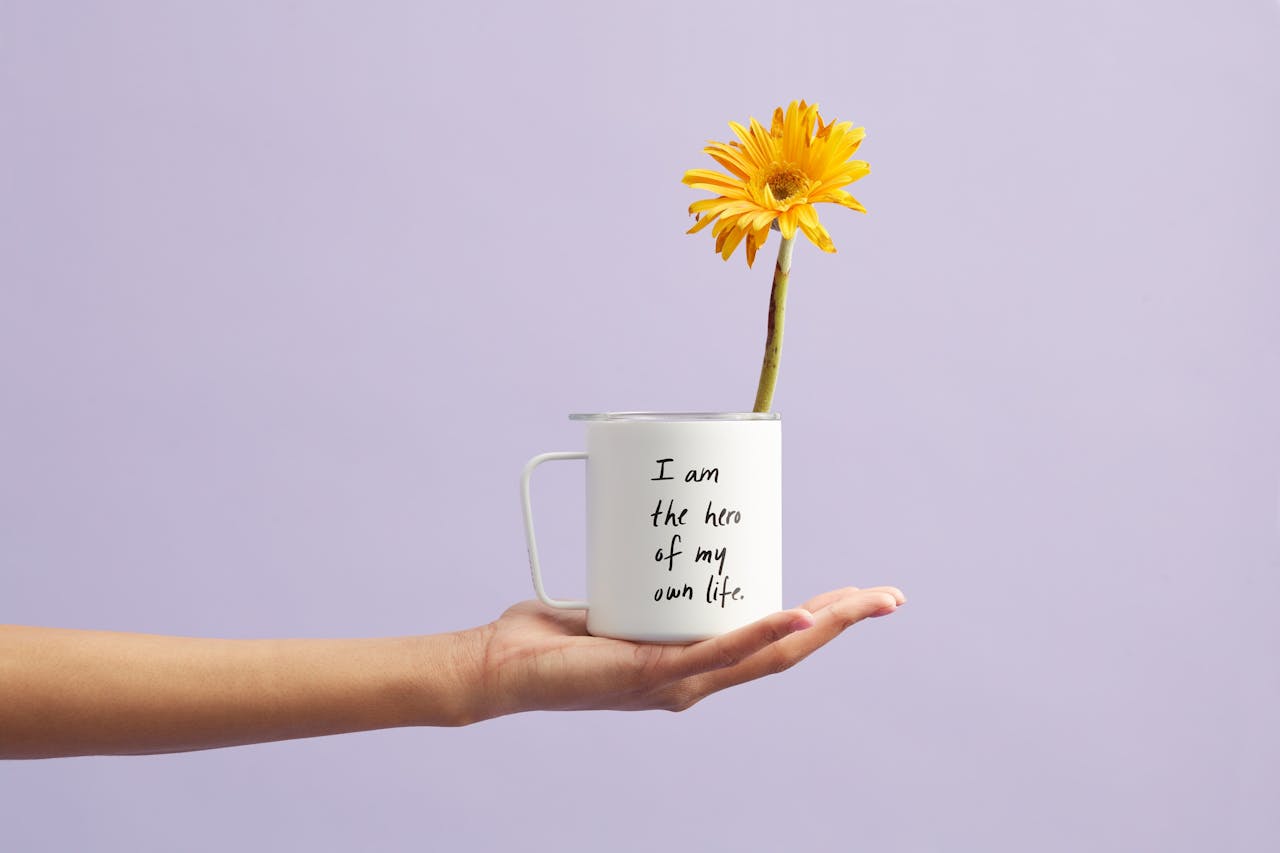 Hand holding a mug with a sunflower and motivational quote, set against a purple background.